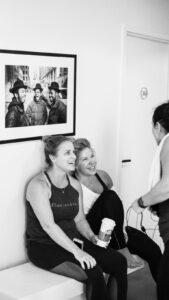 This image displays two women sitting smiling up at a woman standing in front of them. 