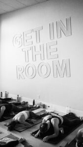This image displays a large sign hung on that wall that says, “Get in the room” with people doing yoga underneath 