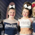 Four smiling cheerleaders stand side by side with their arms around each other, posing in front of a blurred competition backdrop. They wear coordinated navy, white, and silver rhinestone uniforms with "PACK" across the front and large bows in their hair. Three athletes wear long-sleeve tops, while one in the center wears a cropped cheer uniform. The group faces the camera under bright stage lighting, conveying a celebratory team moment.