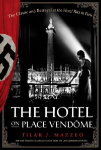 Book cover of The Hotel on Place Vendôme by Tilar J. Mazzeo. A black-and-white nighttime photograph shows a woman in an elegant dress standing on a balcony overlooking Place Vendôme in Paris, with the Vendôme Column illuminated by a dramatic spotlight. A red Nazi flag hangs along the left edge of the cover, contrasting with the monochrome cityscape and emphasizing the book's World War II setting. The subtitle reads, "The Classic and Betrayal at the Hotel Ritz in Paris."