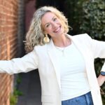 This image displays a smiling woman with shoulder-length curly blonde hair stands outdoors beside a red brick wall, wearing a cream blazer over a white top with blue jeans. She poses with one hand resting on the wall and the other on her hip, while green foliage and a softly blurred walkway create a warm, natural background.