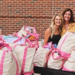 Three women stand smiling behind a large collection of cream-colored tote bags with bright pink handles and trim, arranged on tables outdoors in front of a red brick wall. The bags appear filled with various items, suggesting a donation drive, community event, or organized distribution effort.