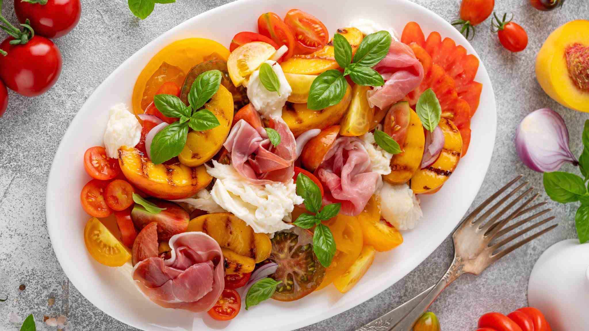 A colorful summer salad with heirloom tomatoes, grilled peaches, fresh mozzarella, prosciutto, red onion, and basil served on a white platter.