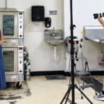 A woman in a navy-blue dress and tan belt poses beside commercial ovens in a kitchen while a man photographs her with a professional camera. Studio lighting and photography equipment are set up between them.
